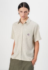 Kortermet, lys beige skjorte med vertikale svarte striper, knappefront, brystlomme og en brodert design på lommen.