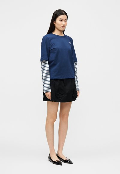 Νεαρή γυναίκα στέκεται, φοράει μπλε σκούρο μπλουζάκι με κοντά μανίκια πάνω από ριγέ μακριά μανίκια, μαύρη φούστα με λουλουδένιες λεπτομέρειες και μαύρα μυτερά παπούτσια μπαλέτου.