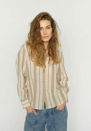 Donna con capelli lunghi e ondulati che indossa una camicia a righe con bottoni e jeans blu, in piedi con le mani in tasca contro uno sfondo semplice.