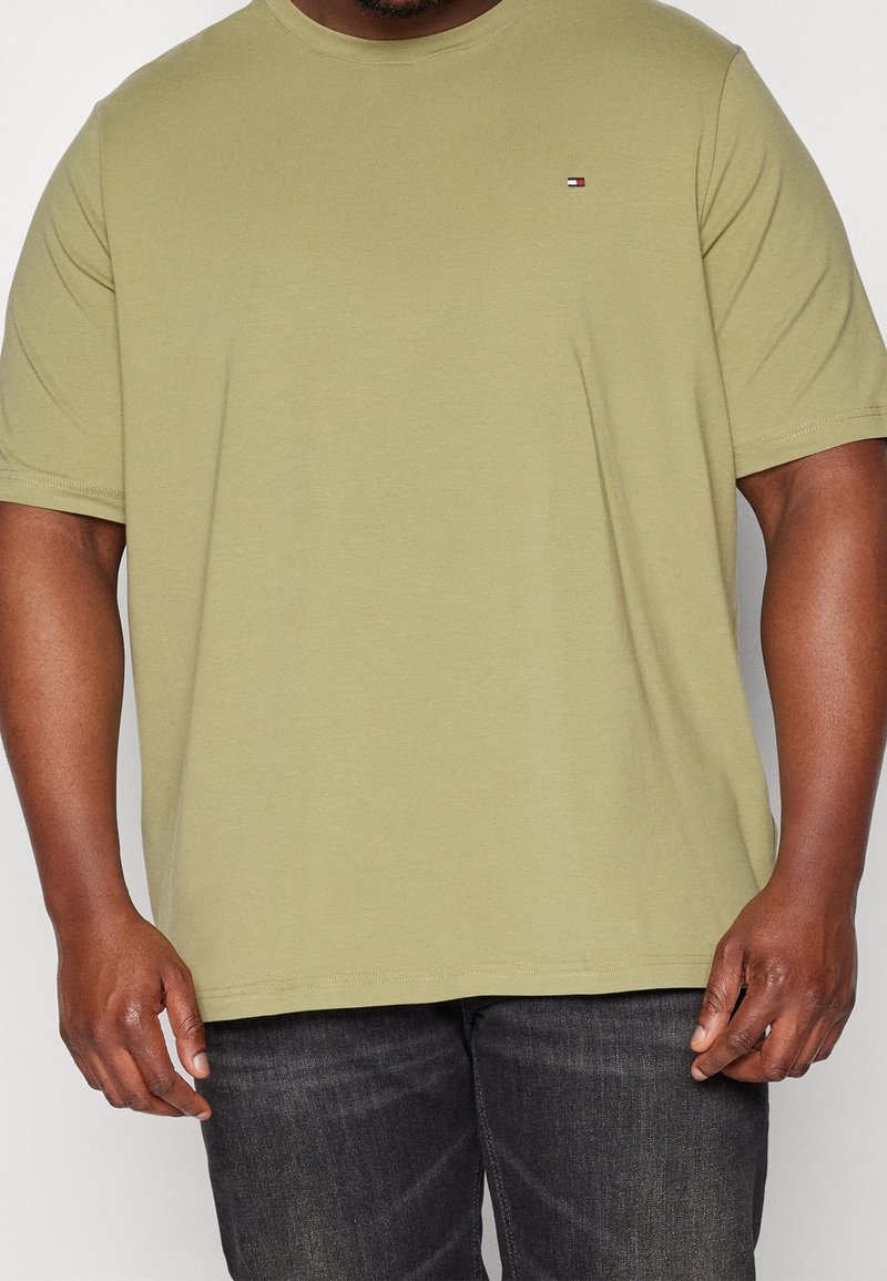 Homme portant un t-shirt à manches courtes vert olive clair avec un petit logo Tommy Hilfiger, assorti à un jean gris foncé.