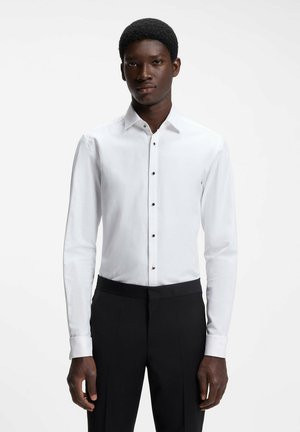 Mężczyzna ubrany w białą koszulę z długim rękawem z czarnymi guzikami i czarne eleganckie spodnie, stojący na jednolitym białym tle.