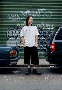 Bela majica s kratkimi rokavi z logotipom Umbro, črne kratke hlače do kolen, črni škornji, stoji med dvema parkiranima avtomobiloma ob grafitnem zidu.