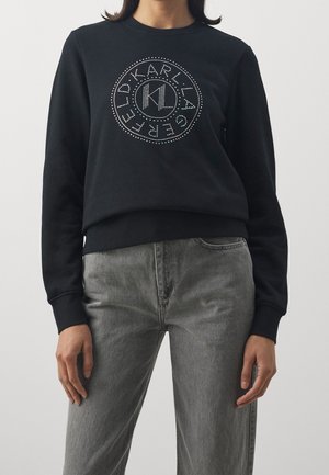 Osoba nosící černou mikinu s kulatým logem a kruhovým puntíkovým vzorem, doplněnou šedými džíny s vysokým pasem.