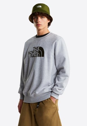 Sweat-shirt gris clair avec logo noir, poignets et col côtelés. Associé à un short marron et un bob vert. Tenue décontractée et sportive.