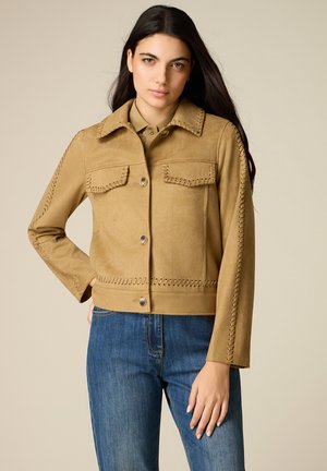 Frau mit langen dunklen Haaren trägt eine hellbraune Wildlederjacke mit gesteppten Details und blaue Jeans, posiert vor neutralem Hintergrund.