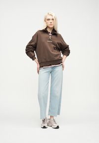 Νεαρό άτομο με μακριά ξανθά μαλλιά που φοράει καφέ φούτερ, ανοιχτό μπλε τζιν και μπεζ αθλητικά παπούτσια, στέκεται μπροστά σε απλό λευκό φόντο.