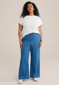 Camiseta blanca de manga enrollada, combinada con pantalones de mezclilla azul de pierna ancha con una cintura ajustable. El calzado incluye zapatillas blancas.