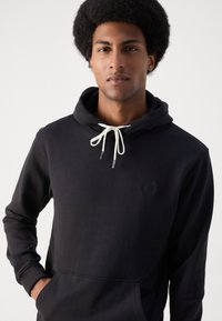 Haut à capuche noir en coton doux, doté d'une poche kangourou, d'un cordon de serrage réglable avec des embouts de couleur claire, et d'une discrète étiquette avec logo.