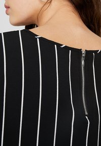 Primo piano della schiena di una donna che indossa un abito nero con strisce bianche verticali e una cerniera argentata visibile all'altezza del colletto.