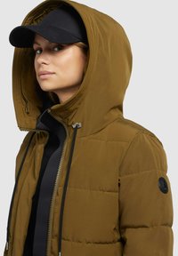 Brown puffer jacket with a large hood, featuring a black zip, drawstrings, and a circular black logo on the sleeve. Worn with a black cap.