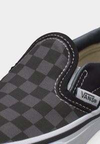 Κοντινή λήψη ενός μαύρου και γκρι καρώ παπουτσιού Vans χωρίς κορδόνια, με λευκές ραφές και λευκή ελαστική σόλα.