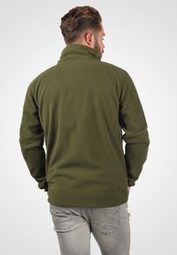 Olive green fleece jacket with high collar, textured fabric, and two vertical seams at the back. Worn with light gray pants.