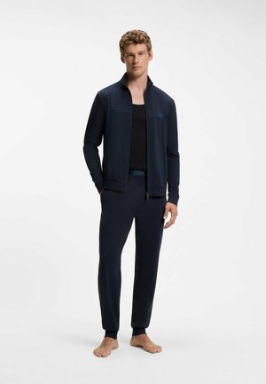 Junger Mann steht barfuß, trägt eine dunkelblaue Reißverschlussjacke, passende Hose und schwarzes ärmelloses Shirt auf weißem Hintergrund.