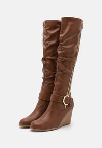 Bottes compensées montantes marron jusqu'au genou avec fermeture éclair latérale et sangle ornée d'une boucle dorée, finition cuir texturée légèrement froissée.