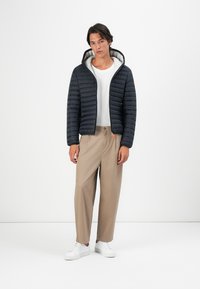 Marineblauwe pufferjack met capuchon, horizontale padding, grijze voering; witte t-shirt; beige broek; witte sneakers.