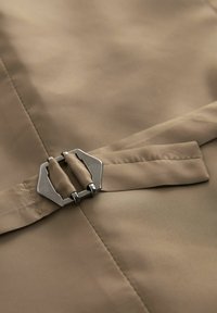 Beige Stoff mit glatter Textur. Verfügt über eine rechteckige Metallbuckle, die an einem gefalteten Riemen befestigt ist. Sauber sichtbare Nähte.