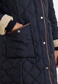 Mornarski podloženi kaput s bež umjetnim krznom na rukavima, smešnim obrubom, pomičnim dugmadi i bočnim džepovima. Teksture s dijamantnim uzorkom. 