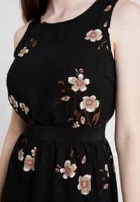 Mustat hihattomat mekko, jossa on beigejä ja ruskeita kukkakuoseja, vartalonmyötäinen malli leveällä mustalla vyötärönauhoilla.
