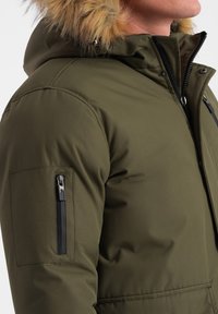 Chaqueta aislante verde oliva con capucha de piel sintética, bolsillo lateral con cremallera y puños de canalé. Textura suave, diseño moderno y detalles de herrajes resistentes.
