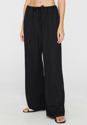 Woman wearing loose black wide-leg pants with drawstring waist and black sandals with gold accents, standing against plain background.