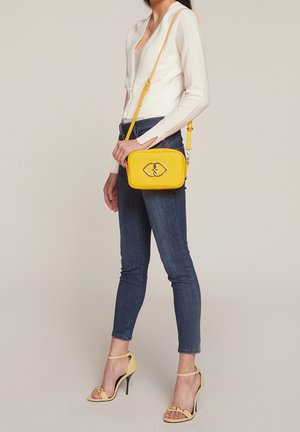 Kobieta ubrana w biały dopasowany top, niebieskie obcisłe dżinsy, żółte sandały na wysokim obcasie oraz z żółtą torebką na ramię z metalową klamrą.