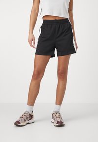 Black athletic shorts with an elastic waistband, side stripes, and a loose fit. Worn with a cropped white top and patterned sneakers.