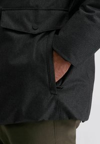 Schwarze, strukturierte Jacke mit Karomuster, ausgestattet mit einer Seitentasche und einem subtilen Glanz. Das Material wirkt glatt und wasserabweisend.