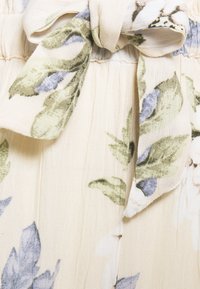 Látka s květinovým vzorem v béžové barvě, zdobená zelenými a modrými listy. Obsahuje zavazovací pas s třepeným okrajem pro texturu.