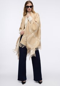 Sciarpa beige con bordi sfrangiati, caratterizzata da un motivo a logo beige chiaro ripetuto. Modello che indossa occhiali da sole neri e jeans scuri a zampa d'elefante.