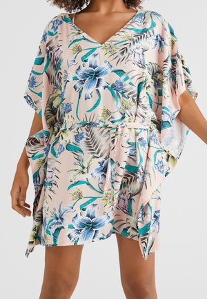 Femme portant une robe ample à motif floral avec des fleurs bleues, vertes et jaunes sur fond rose clair et un décolleté en V.