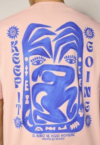 Camiseta de algodón de color rosa claro con un gran diseño gráfico azul que presenta rostros abstractos, texto y elementos estilizados. Corte relajado, cuello redondo.