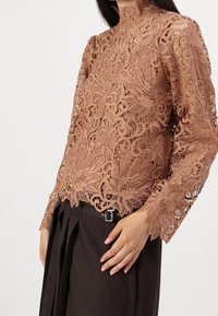 Haut en dentelle à manches longues de couleur brun clair avec des motifs floraux détaillés, col montant et texture transparente, assorti à une jupe marron foncé.