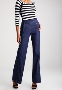 Pantalones de mezclilla de pierna ancha de tiro alto en un azul oscuro, con una textura suave, que presentan llamativos botones en la parte frontal y se complementan con tacones negros con tirantes.