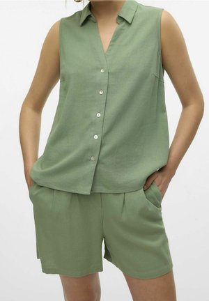 Chemise sans manches vert clair à boutons, associée à un short vert assorti, portée les mains dans les poches devant un fond uni.