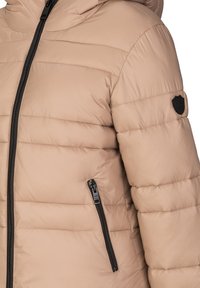 Beige Pufferjacke mit horizontaler Naht, hohem Kragen, seitlichem Reißverschluss und einem schwarzen Logo-Patch am Ärmel.