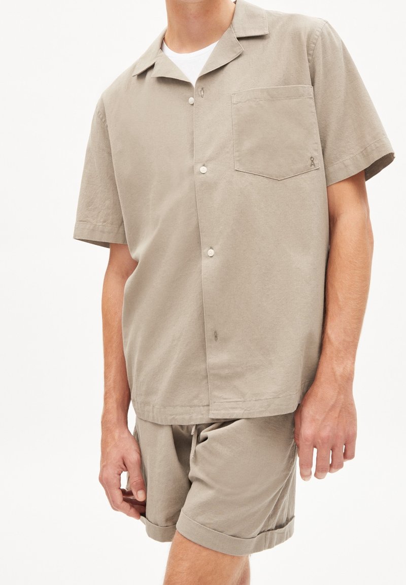 Camicetta a maniche corte di un leggero beige con bottoni e una tasca sul petto, abbinata a pantaloni corti coordinati, caratterizzata da un design semplice e casual con orli risvoltati.