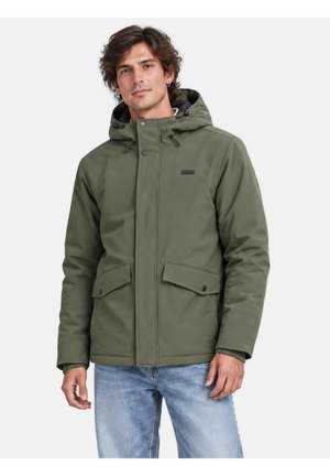 Homme aux cheveux bouclés foncés portant une veste d’hiver à capuche couleur olive et un jean bleu clair, debout sur un fond blanc.