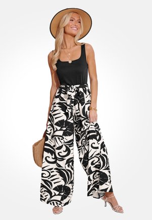 Blonde Frau mit schwarzem Tanktop, weit geschnittenen schwarz-weiß gemusterten Hose mit gebundener Taille, breitkrempigem Strohhut und weißen Riemchen-Absatzschuhen.