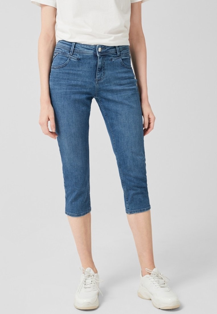 Pantalones capri de mezclilla en azul medio con un diseño ajustado, estilo de cinco bolsillos y un dobladillo justo por debajo de la rodilla. Llevados con zapatillas blancas.