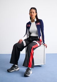 Marinblå och vit dragkedjegenomgående sportjacka med röd text, i kombination med svarta byxor med röda och vita detaljer samt svarta sneakers.