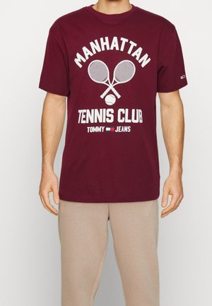 Homme portant un t-shirt bordeaux avec un motif blanc sur le thème du tennis indiquant « Manhattan Tennis Club Tommy Jeans » et un pantalon beige.
