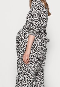 Robe noire à fleurs avec des fleurs blanches et roses, manches smockées, vue de côté montrant une coupe ample et une taille froncée, texture de tissu douce.