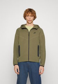 Olive green zip-up hoodie with a hood, black zippers, and two side pockets. Features textured fabric and contrasting seams.