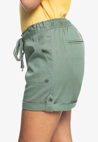 Vue latérale d'une personne portant un short vert à cordon de serrage avec ourlet roulé et une chemise jaune, mettant en avant les détails et la coupe du short.