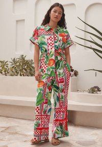 Skok kombinezon cvetličnega potiska z živahnimi odtenki zelene, rdeče in rumene, z ovijajočim dizajnom, kratkimi rokavi in širokimi hlačnicami.