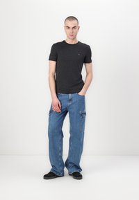Dunkelgraues T-Shirt kombiniert mit hellblauen Cargo-Jeans, die seitliche Taschen haben. Das Model trägt schwarze Schuhe und steht vor einem schlichten Hintergrund.