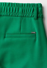 Pantalons en tissu vert avec une taille élastique et une poche arrière. Caractéristiques : coupe ajustée et petite étiquette de marque en métal sur la poche.