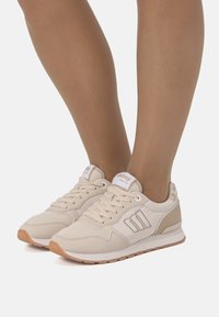 Zapatillas deportivas beige con empeine texturizado, cordones blancos y suela de goma. Presentan un logo en la lengüeta y tres franjas en el lateral.