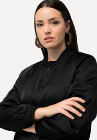 Giubbotto bomber nero con zip frontale, colletto e polsini a costine; texture liscia; indossato da una persona con le braccia incrociate.