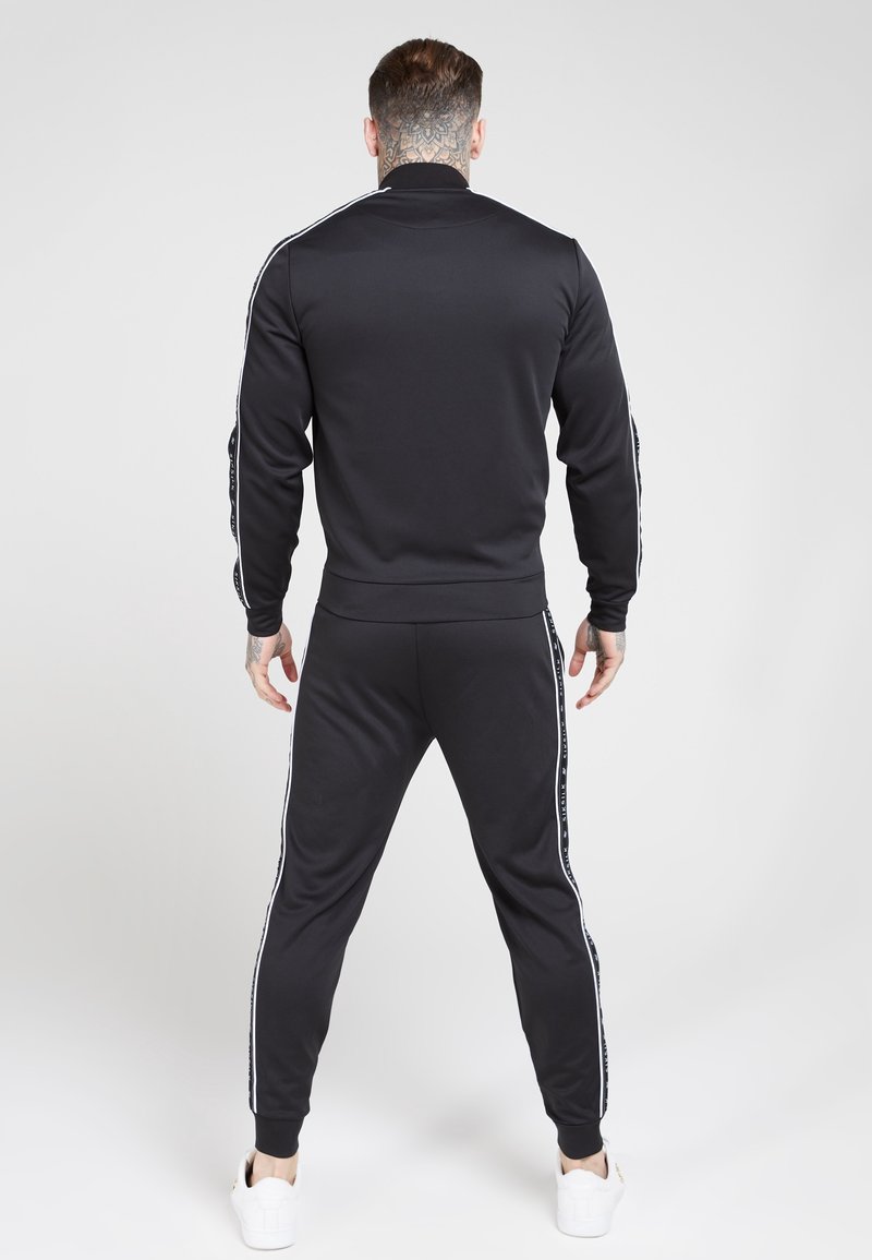 Black tracksuit featuring a zip-up jacket and tapered pants. Both have white side stripes. Material appears smooth. Model stands facing away.